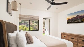 A bedroom with a bed, a ceiling fan, a window with a view of greenery, and a wooden dresser. - Hillside Haven | Wellness Spa, Pickleball, Games (Sedona)