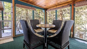 Un espace extérieur aménagé, abrité et doté de chaises en osier et d’une table ronde, entouré de portes et de fenêtres en verre offrant une vue sur les arbres.