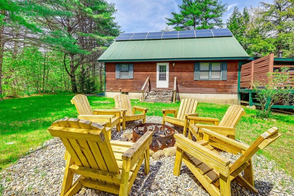A cabin with a green roof, surrounded by a fire pit with chairs, set in a forested area.