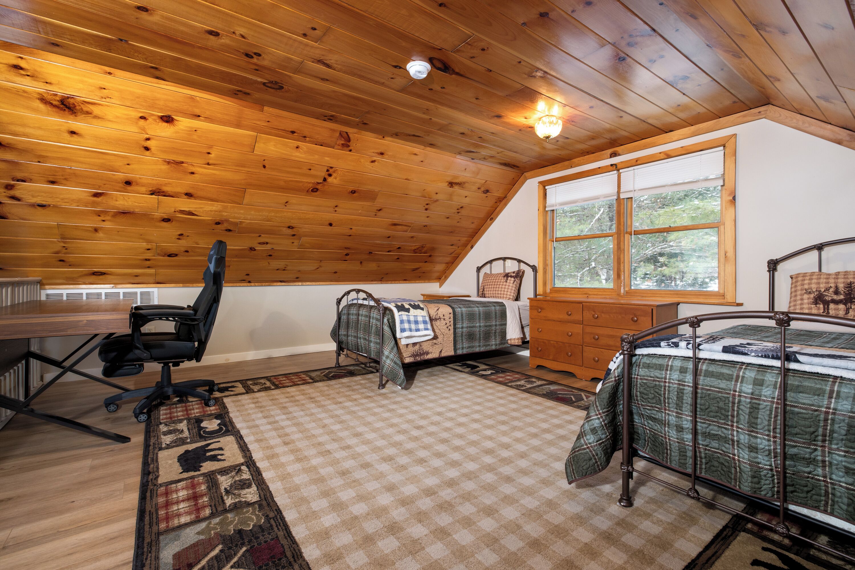 A cozy cabin room with a wooden ceiling, a desk with a chair, two beds with checkered bedding, a dresser, and a window with a view of trees.