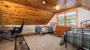 A cozy cabin room with a wooden ceiling, a desk with a chair, two beds with checkered bedding, a dresser, and a window with a view of trees.