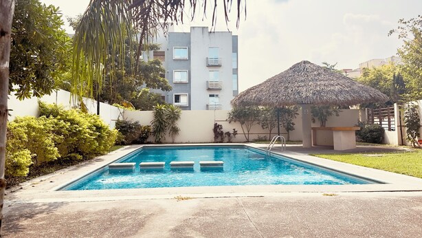 Piscina com degraus, rodeada por vegetação e um edifício com telhado de palha.