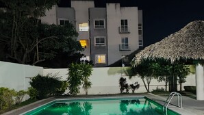 Uma piscina com espreguiçadeiras, uma estrutura com telhado de palha e um edifício com várias janelas ao fundo.