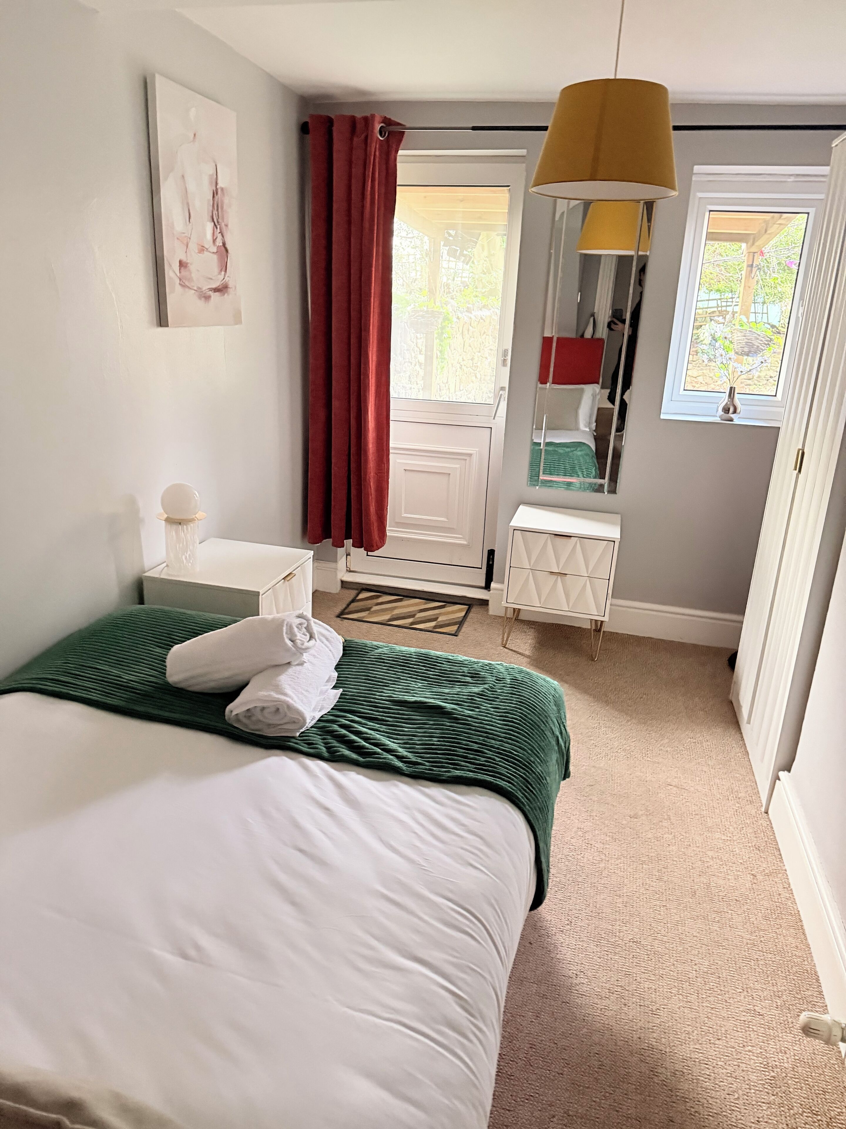 A neatly made bed with a green blanket and white pillows, a white bedside table, a mirror, a window with a view of greenery, and a door leading outside.