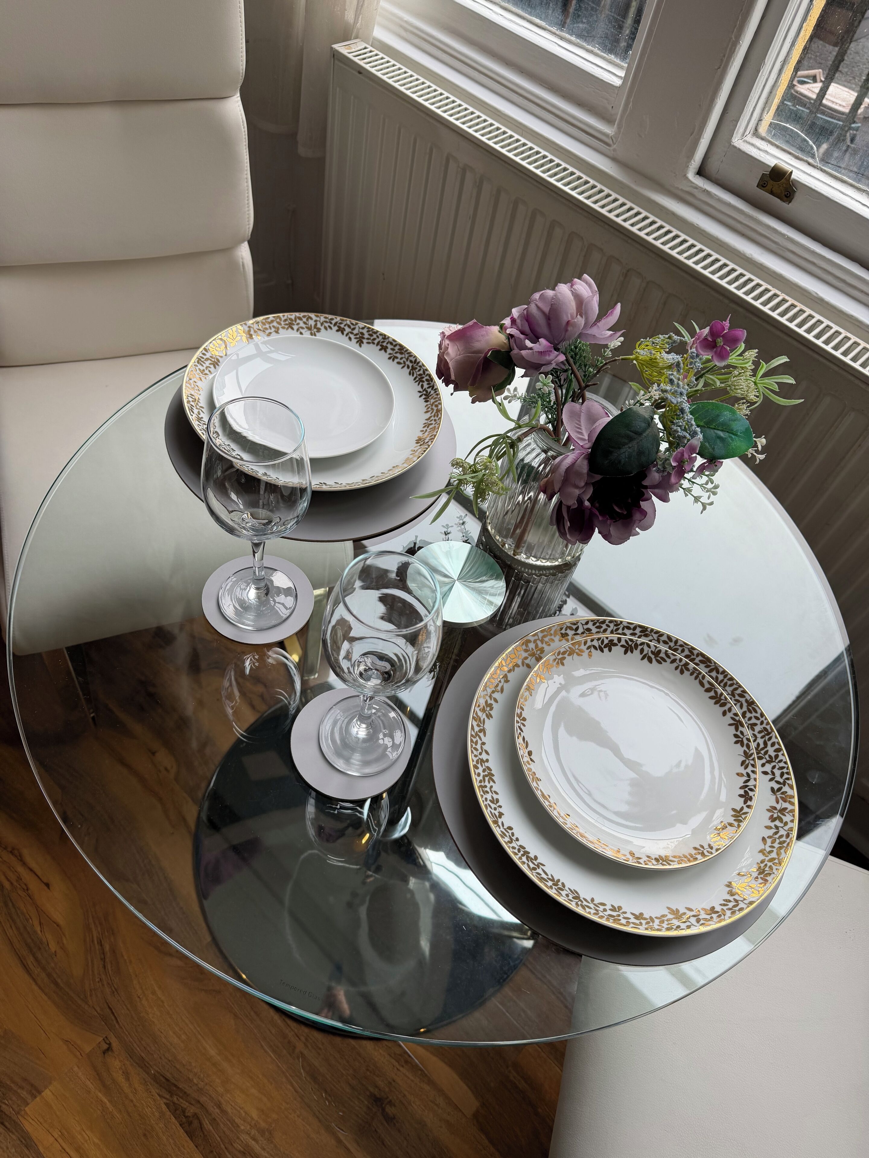Een ronde glazen tafel gedekt met witte borden en gouden accenten, een vaas met bloemen en wijnglazen.