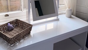 Un escritorio blanco con un espejo, una cesta de mimbre y un difusor de caña.