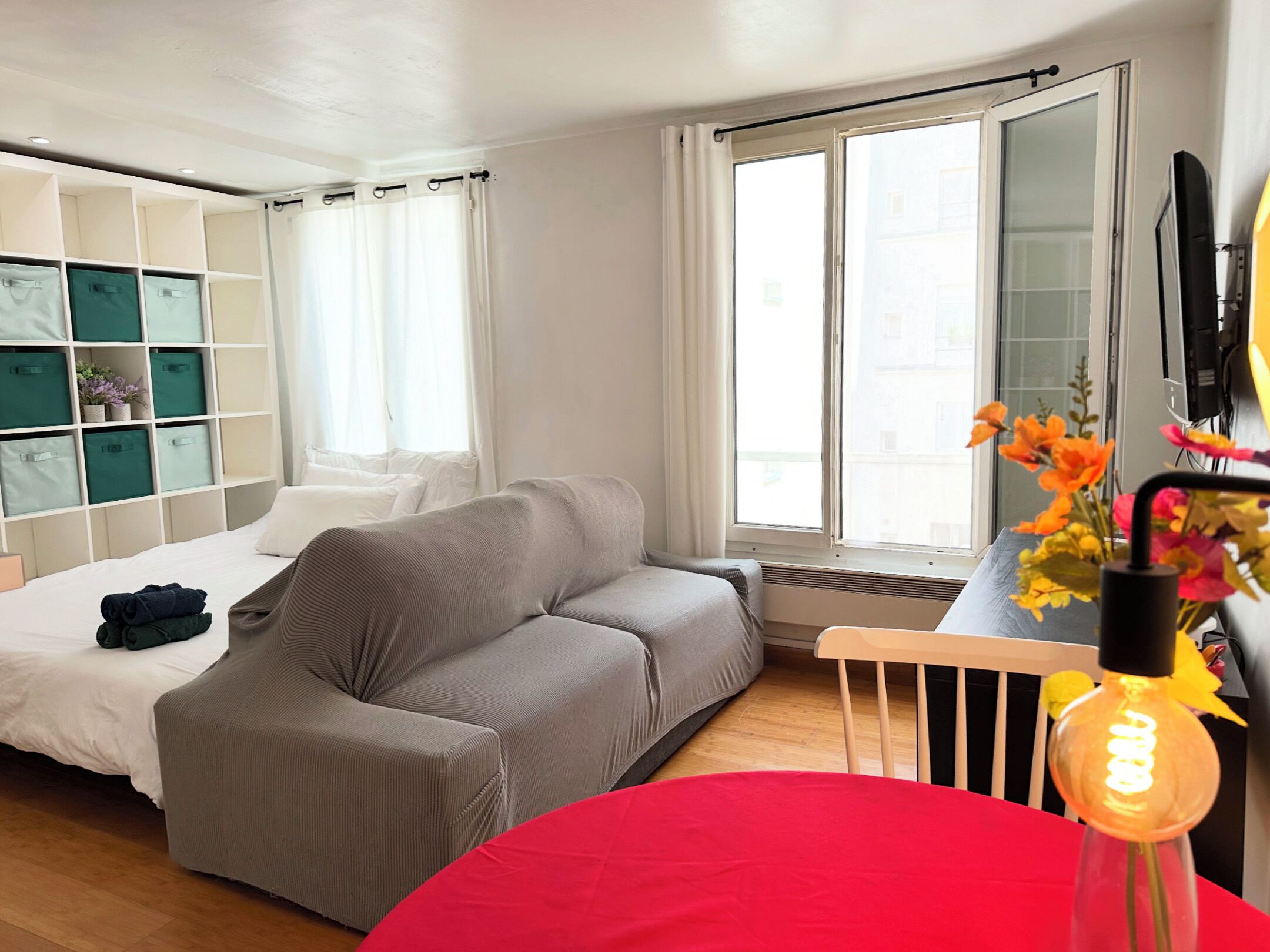 Een moderne slaapkamer met een grijze bank, een rode fauteuil, een bed met witte beddengoed, een inbouwkleerkast met groene opbergdozen, en een groot raam met gordijnen.