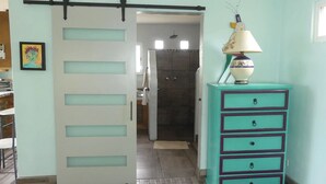A room with a sliding door leading to a bathroom, a teal dresser with drawers, and a lamp on top. - Siren Casita - Incredible Ocean Views, A/C & Heated Saltwater Pool (Todos Santos)