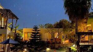 A well-lit outdoor area with a pool, palm trees, and a building with a yellow facade. - Siren Casita - Incredible Ocean Views, A/C & Heated Saltwater Pool (Todos Santos)