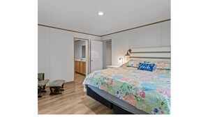 Un dormitorio con cama, mesita de noche, banqueta y puerta que da a un baño.