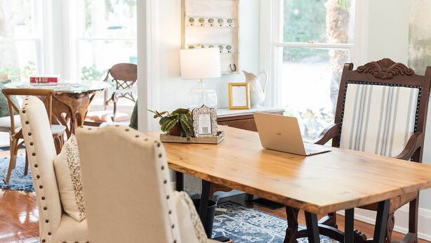 A wooden dining table with a laptop, surrounded by upholstered chairs. A decorative wall piece and a small potted plant are on the table.