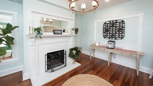 A room with a white fireplace, a chandelier, a console table with a coffee maker, and a decorative wall piece.