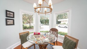 A dining room with a round glass table, wooden chairs, a chandelier, and large windows with a view of the street and greenery.