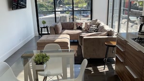 En moderne stue med et glasbord, en beige sofa og et køkken med træskabe.