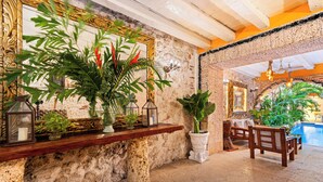 Pared de piedra con repisa de madera que sostiene plantas y un farol. Se ve un espejo grande con marco dorado, un banco de madera y una zona de piscina al fondo.