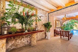 A stone wall with a wooden shelf holding plants and a lantern. There is a large mirror with a gold frame, a wooden bench, and a pool area visible in the background. - 15BR Mansion in the Walled City / Staff Included (Cartagena de Indias)