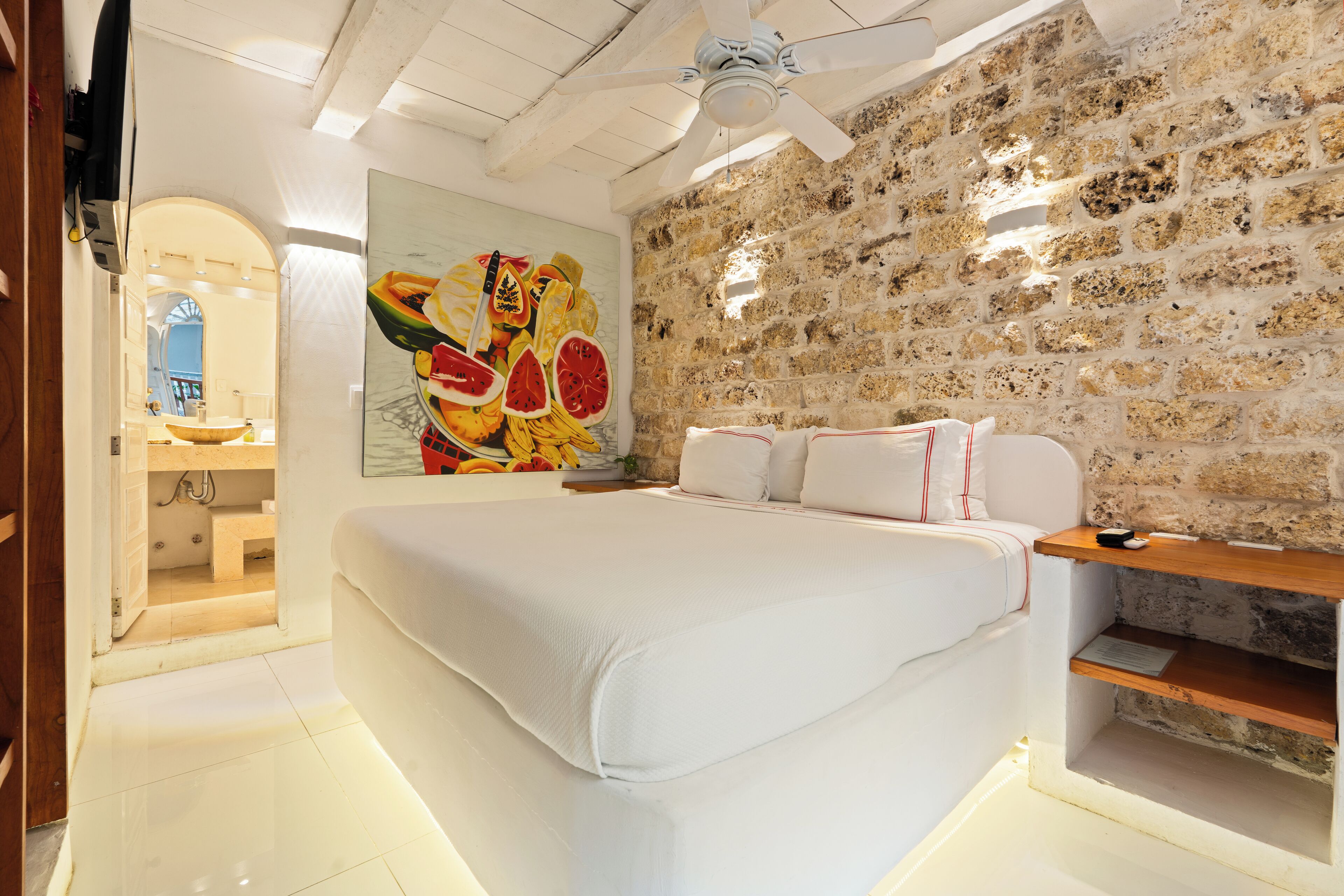 Ein Schlafzimmer mit einem großen Bett, einer Steinsäule, einem Deckenventilator und einem Gemälde mit Früchten.
