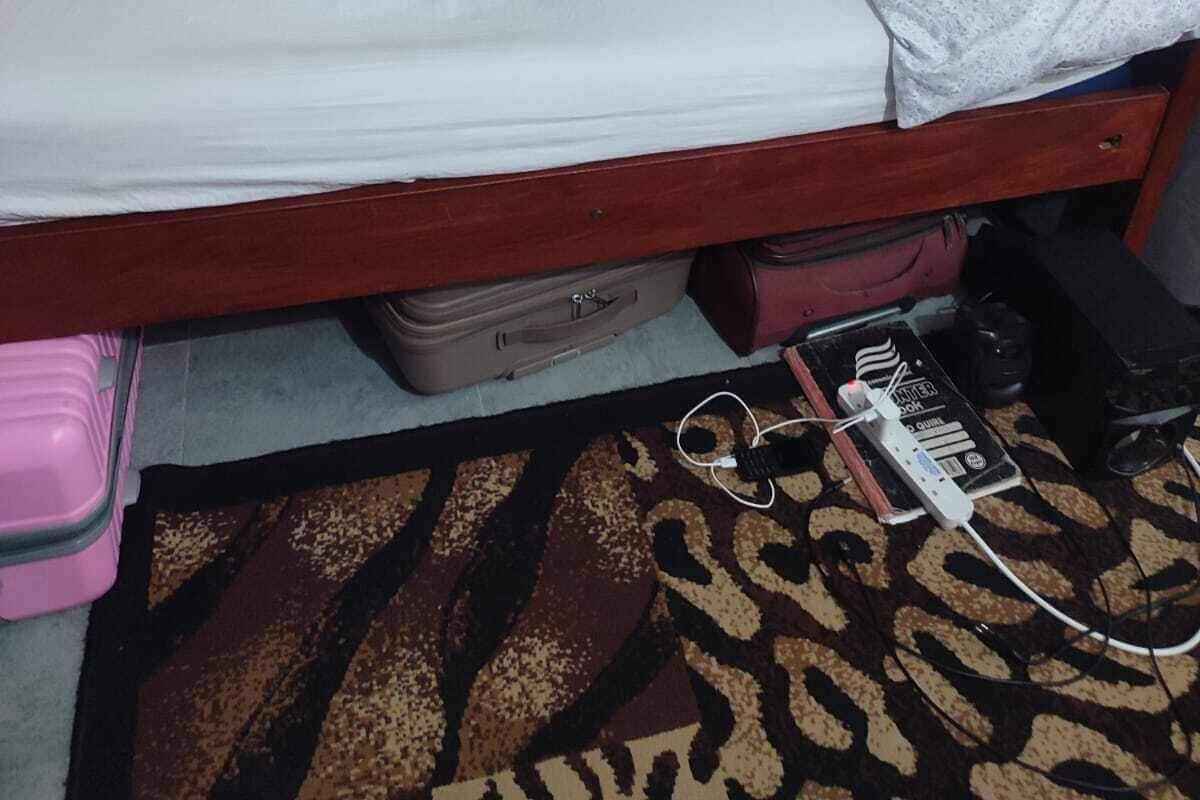 A single bed with a white mattress and a grey blanket, a wooden headboard, a small suitcase under the bed, and a power strip on the floor.