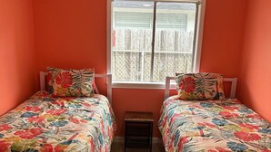 Una habitación con dos camas, una ventana que da a una valla, un ventilador de techo y una mesita pequeña.