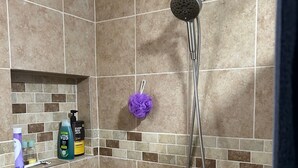 Un área de ducha con cabezal de ducha de mano, un estropajo morado y artículos de aseo en un estante.