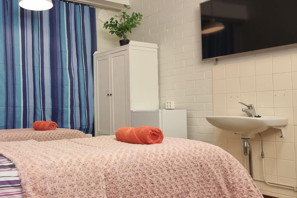 Hotelkamer met twee bedden, gestreept beddengoed, een groot raam met blauwe gordijnen, een witte kast, een potplant en een tv aan de muur.