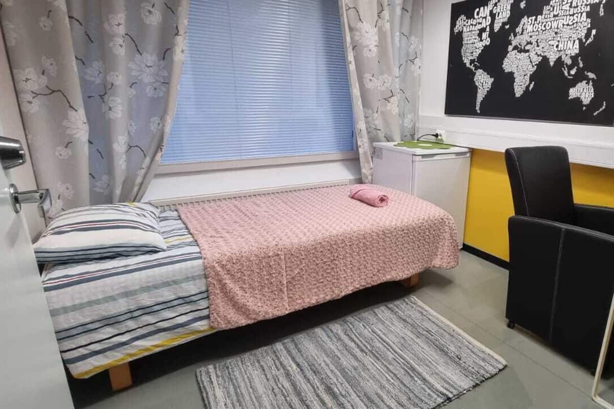 Een kleine kamer met een bed, een bureau en een stoel. Aan de muur hangt een wereldkaart en er is een raam met gordijnen.