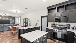 A modern kitchen with dark cabinetry, stainless steel appliances, and a herringbone wood floor. - 5-BD Getaway w/ Pool, Patio, Firepit | Mins to DT (Dallas)