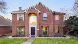 A two-story brick house with a front entrance, large windows, and a well-maintained lawn. - 5-BD Getaway w/ Pool, Patio, Firepit | Mins to DT (Dallas)