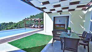 Ein Poolbereich mit Esstisch und Stühlen, einer Pergola mit Liegestühlen sowie Blick auf einen Swimmingpool und Grünflächen.