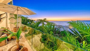A balcony with a view of the ocean at sunset, surrounded by tropical plants and a large umbrella. - New 4BR Luxury Condo at Indah (Puerto Vallarta)