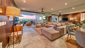 A spacious living room with a large sofa, a coffee table, and a TV. There is a large window with a view of the mountains. - New 4BR Luxury Condo at Indah (Puerto Vallarta)