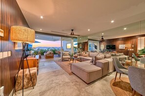 Ein geräumiges Wohnzimmer mit einer großen Couch, einem Couchtisch und einem Fernseher. Ein großes Fenster bietet Blick auf die Berge.