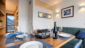 Uma área de refeições com uma mesa posta para dois, uma garrafa de vinho e vista para as montanhas pela janela.