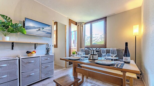 A modern kitchen with a wooden table set for dining, a wine bottle, and a large window with a view of snow-capped mountains. - Alpine Ski-In/Out w/MountainView (Val Thorens)