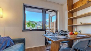 Ein Essbereich mit einem Holztisch, gedeckt für zwei Personen, mit Blick auf Berge durch ein Fenster und einer gemütlichen Sitzecke mit einer Couch und Kissen.