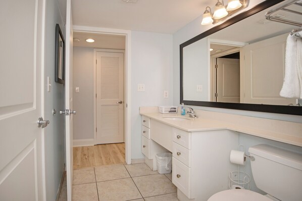 Un baño con un lavamanos blanco, un espejo grande, un inodoro y una puerta que da a otra habitación.