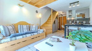 A cozy living room with a wooden sofa, a white coffee table, and a plant. A kitchen with a bar and a staircase leading to another level is visible in the background. - Mountain Hideaway near the Slope (Val Thorens)