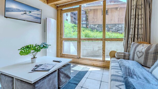 A cozy room with a wooden ceiling, a large window with curtains, a sofa with patterned cushions, a small table with a plant, and a framed picture on the wall. - Mountain Hideaway near the Slope (Val Thorens)