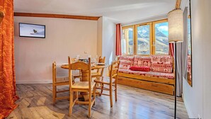 Quarto com mesa de jantar e cadeiras de madeira, sofá com almofadas estampadas, janela grande com vista para as montanhas e televisão fixada na parede.