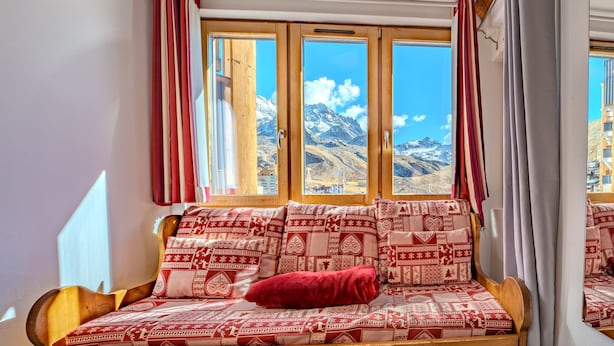 Um sofá de madeira com almofadas estampadas e um travesseiro vermelho, posicionado em frente a uma janela com vista para as montanhas.