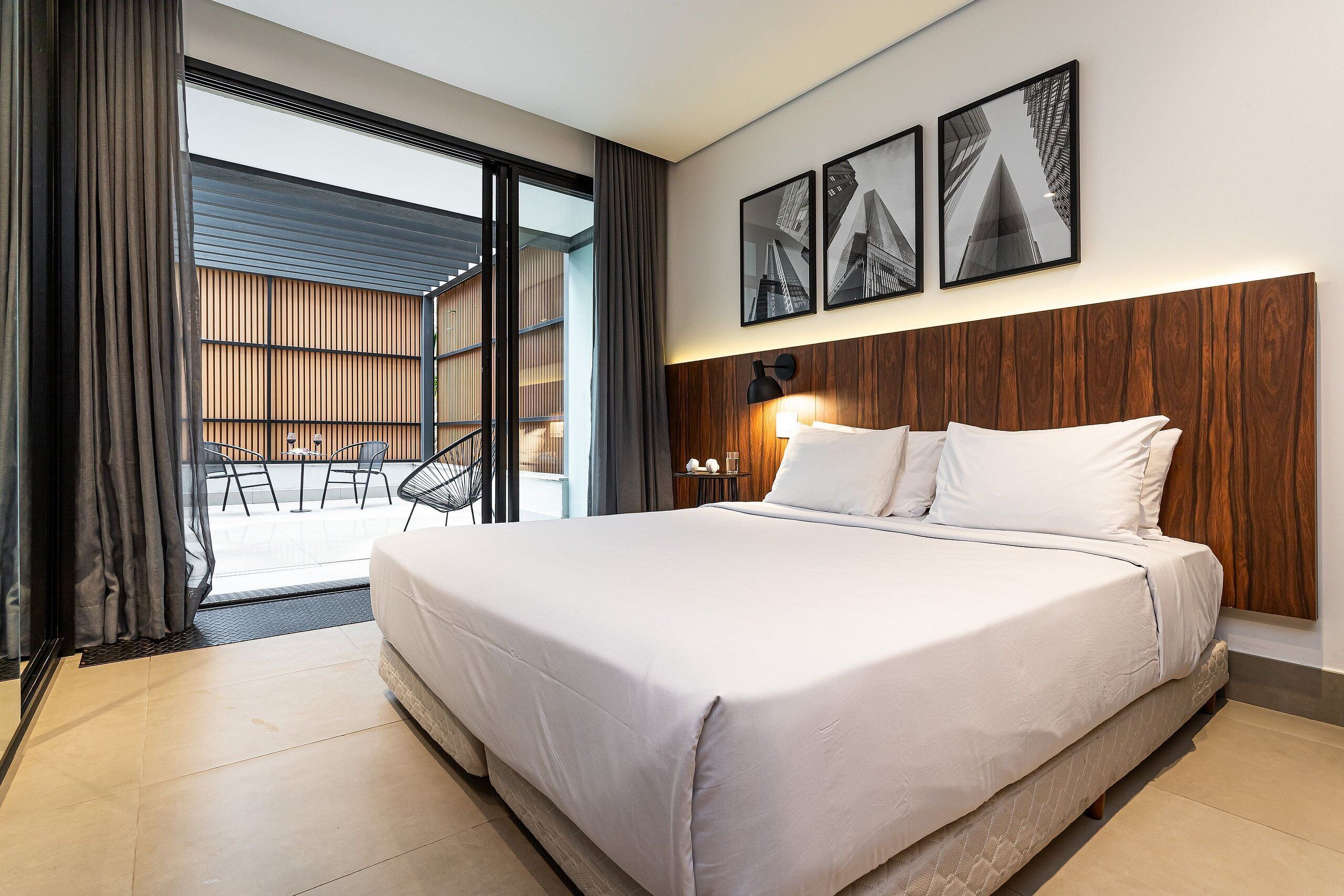 A modern hotel room with a large bed, a wooden headboard, and a view of the city through floor-to-ceiling windows.