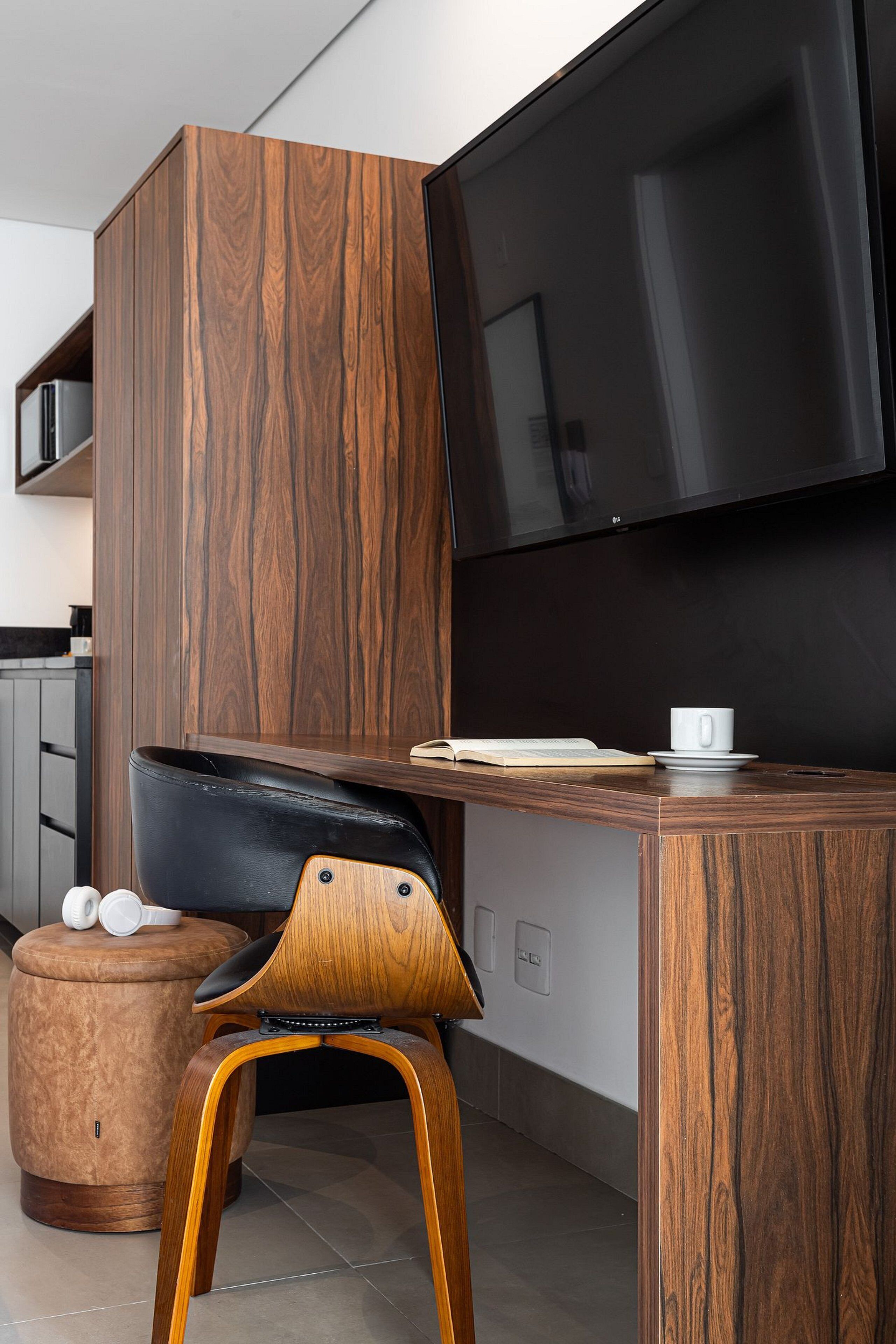Une chambre moderne avec un bureau en bois, une chaise noire, un téléviseur fixé au mur et un petit tabouret sur lequel est posée une tasse.