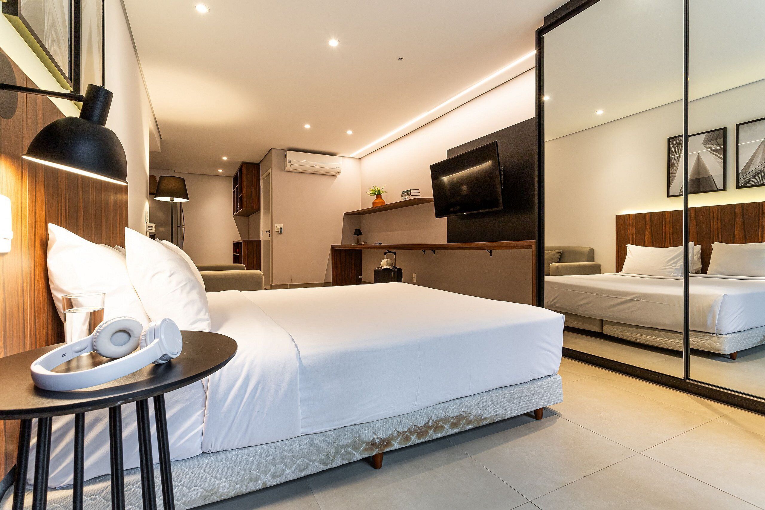 A modern hotel room with a bed, bedside table, headphones, a TV, and a mirror.