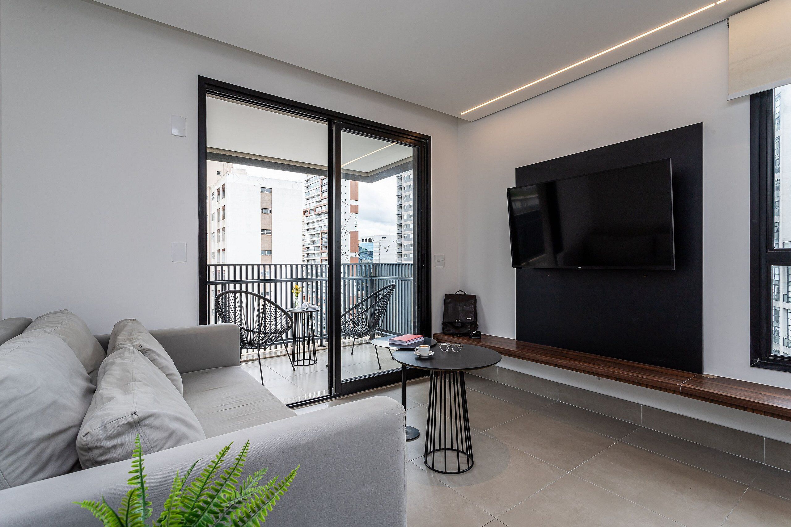 A modern living room with a sofa, a small table, a TV mounted on the wall, and a balcony with chairs.