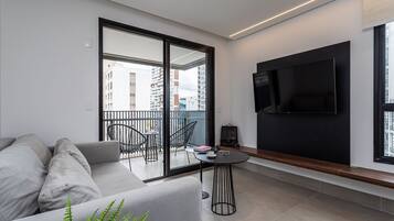 A modern living room with a sofa, a small table, a TV mounted on the wall, and a balcony with chairs.