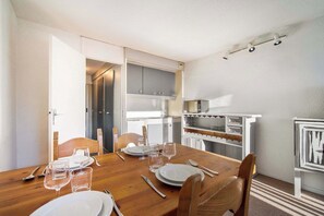 Uma sala de jantar com mesa de madeira posta para quatro pessoas, cozinha com armários brancos e uma pequena geladeira.