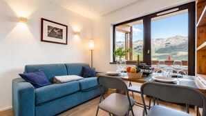 A modern living room with a blue sofa, a dining table set with glasses and a fruit bowl, and a large window offering a mountain view. - Sunny Ski-In/Out Mountain Views (Val Thorens)