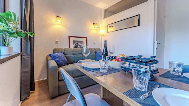 A dining area with a wooden table set for two, two chairs, a sofa, and a potted plant. - Ski Side Nook in Town Center View (Val Thorens)