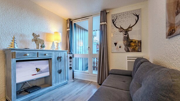 A cozy living room with a grey sofa, a wooden cabinet with a TV, and a deer artwork on the wall. - Slopeside Apt w/ Balcony & Views (Val Thorens)