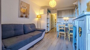 A modern living room with a grey sofa, a dining area with wooden chairs, a kitchen with white cabinets, and a ceiling light fixture. - Slopeside Apt w/ Balcony & Views (Val Thorens)